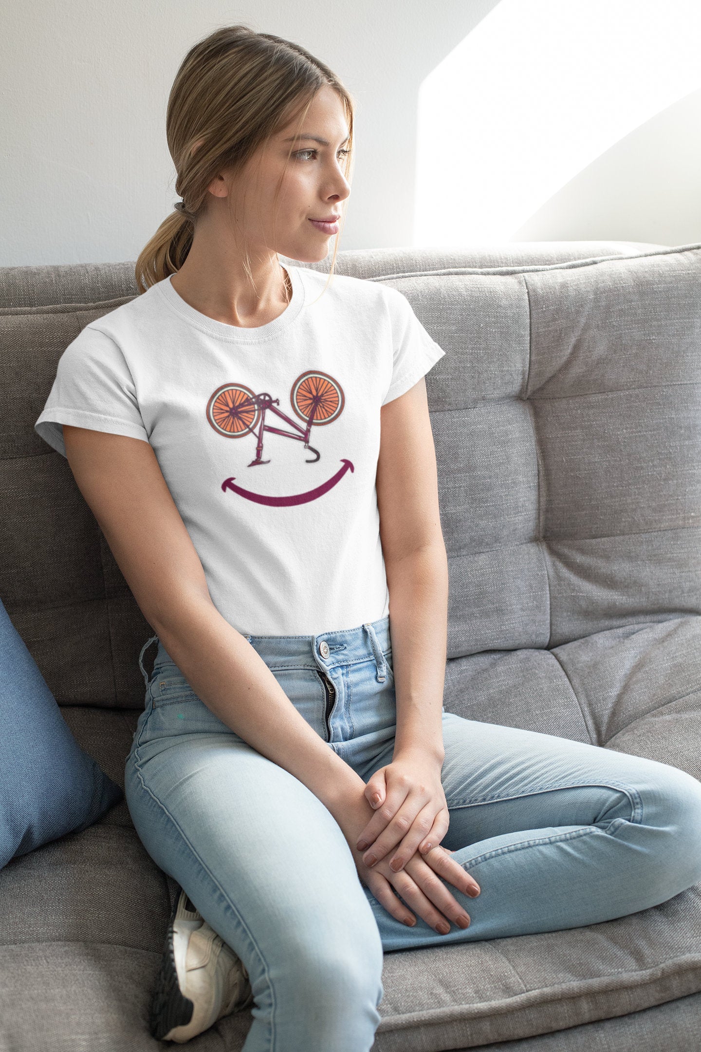 Frau sitzt entspannt auf dem Sofa mit weißem T-Shirt im Smiley-Fahrrad-Design – gemütlicher Lifestyle-Mockup für Radliebhaberinnen