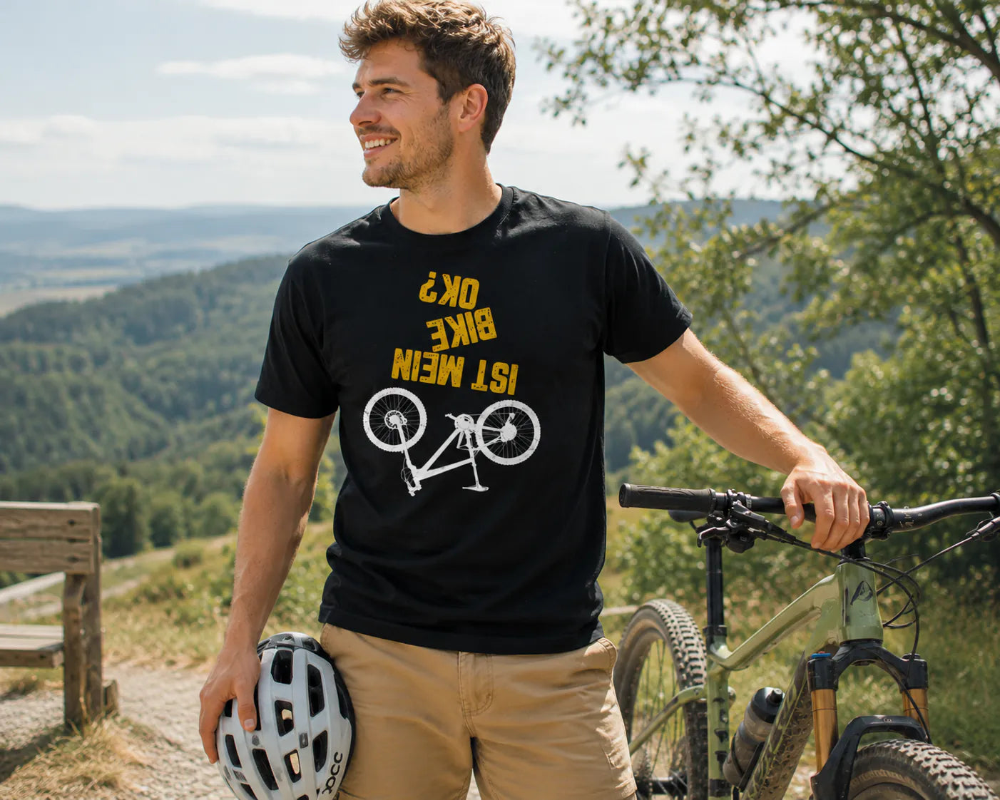 Junger Mann trägt das „Ist mein Bike okay?“ Fahrrad T-Shirt neben einem Mountainbike und hält einen Helm in der Hand