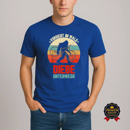 Model trägt blaues Fahrrad-T-Shirt mit Retro-Motiv ‚Vorsicht im Wald – Diebe unterwegs‘ und Bigfoot-Silhouette mit Fahrrad.
